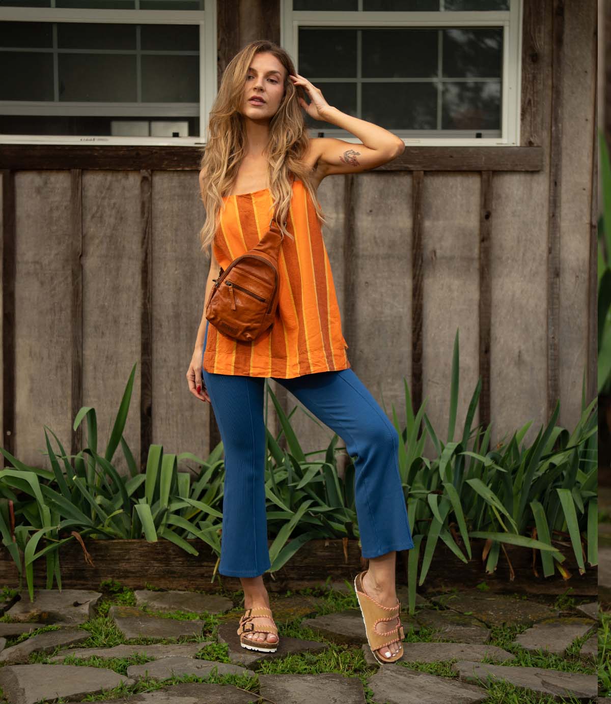 
                  
                    A woman stands on a stone path before a wooden building, wearing an orange striped top, blue pants, tan Roan Mode sandals with cork footbed, and a brown crossbody bag.
                  
                