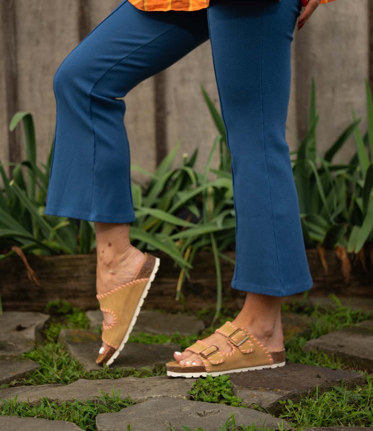 A person in Roan’s Mode blue cropped pants and tan sandals with adjustable buckles stands on stone steps, with green plants in the background.