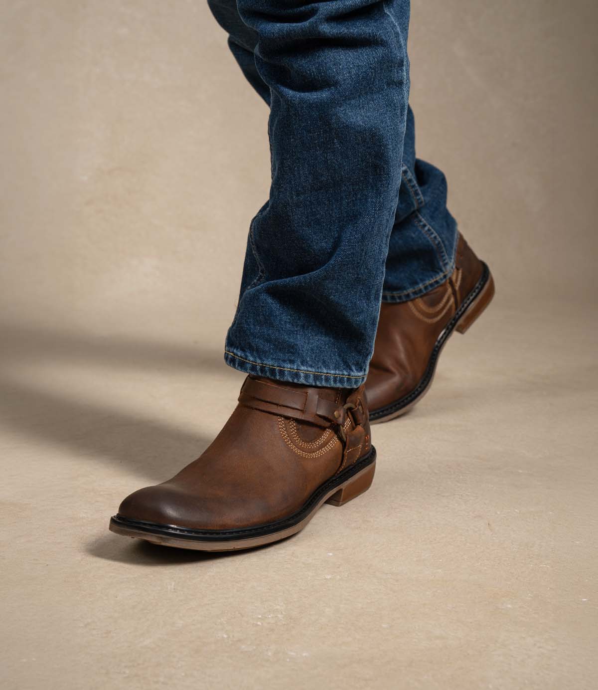 
                  
                    A person wearing blue jeans and a tan Roan Native II leather boot featuring a decorative harness and cushioned insole, walking on a light-colored surface.
                  
                