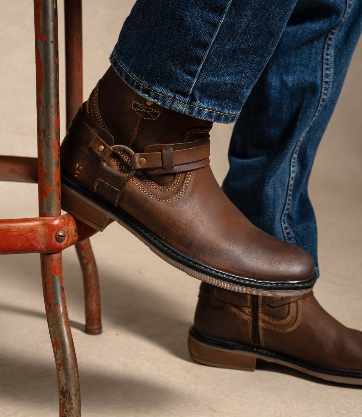 
                  
                    A person wearing blue jeans and the Roan Native II tan Greenland leather boots, featuring a decorative harness and cushioned insole, rests a foot on a rusty metal stool.
                  
                