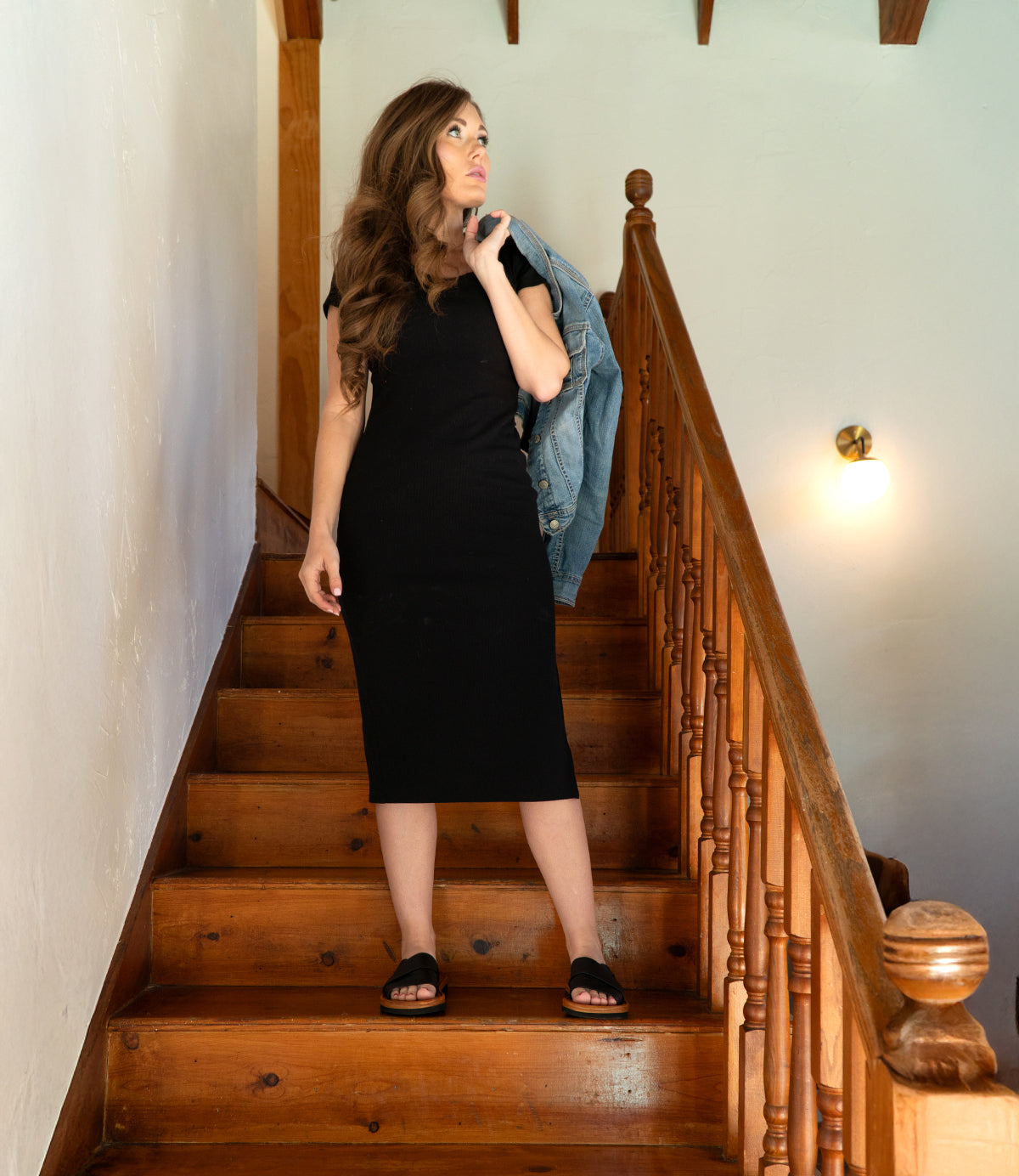 
                  
                    A woman in a black dress stands on a wooden staircase, holding a denim jacket and looking to the side, embodying effortless summer style with her chic Roan Nosy slip-on sandals.
                  
                