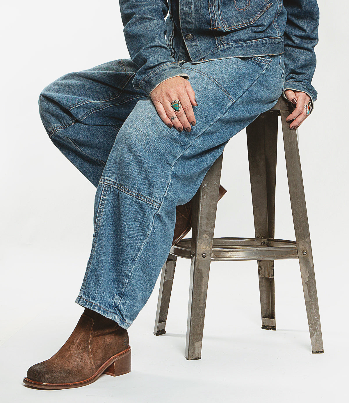 A person dressed in a denim outfit is seated on a metal stool, wearing the Roan "Notice" brown boots featuring a suede upper and square toe, complemented by rings.