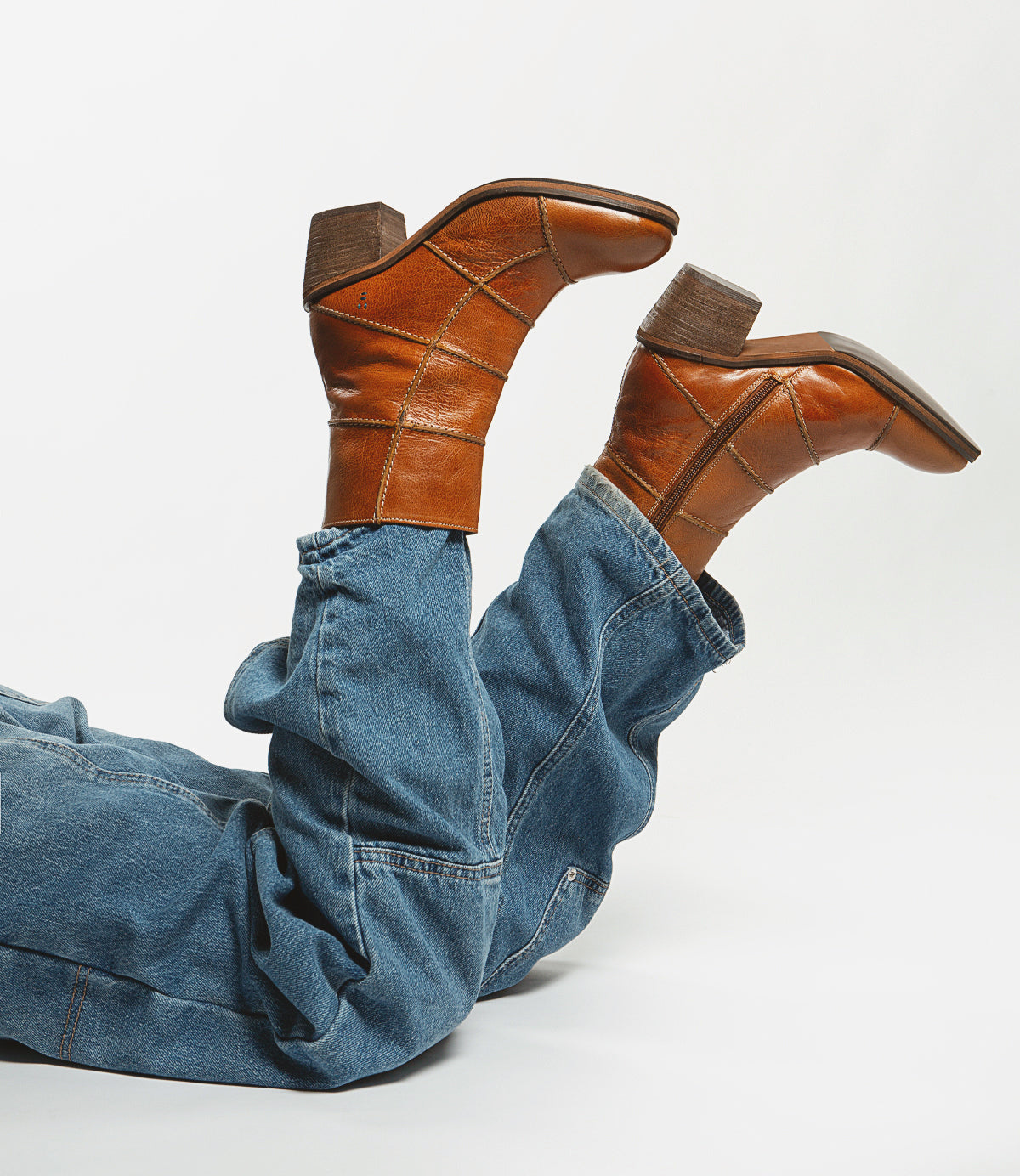A person reclines with their legs up, highlighting the Roan Often blue jeans and brown leather boots with wooden heels—an impactful statement piece that exudes fashion-forward style.
