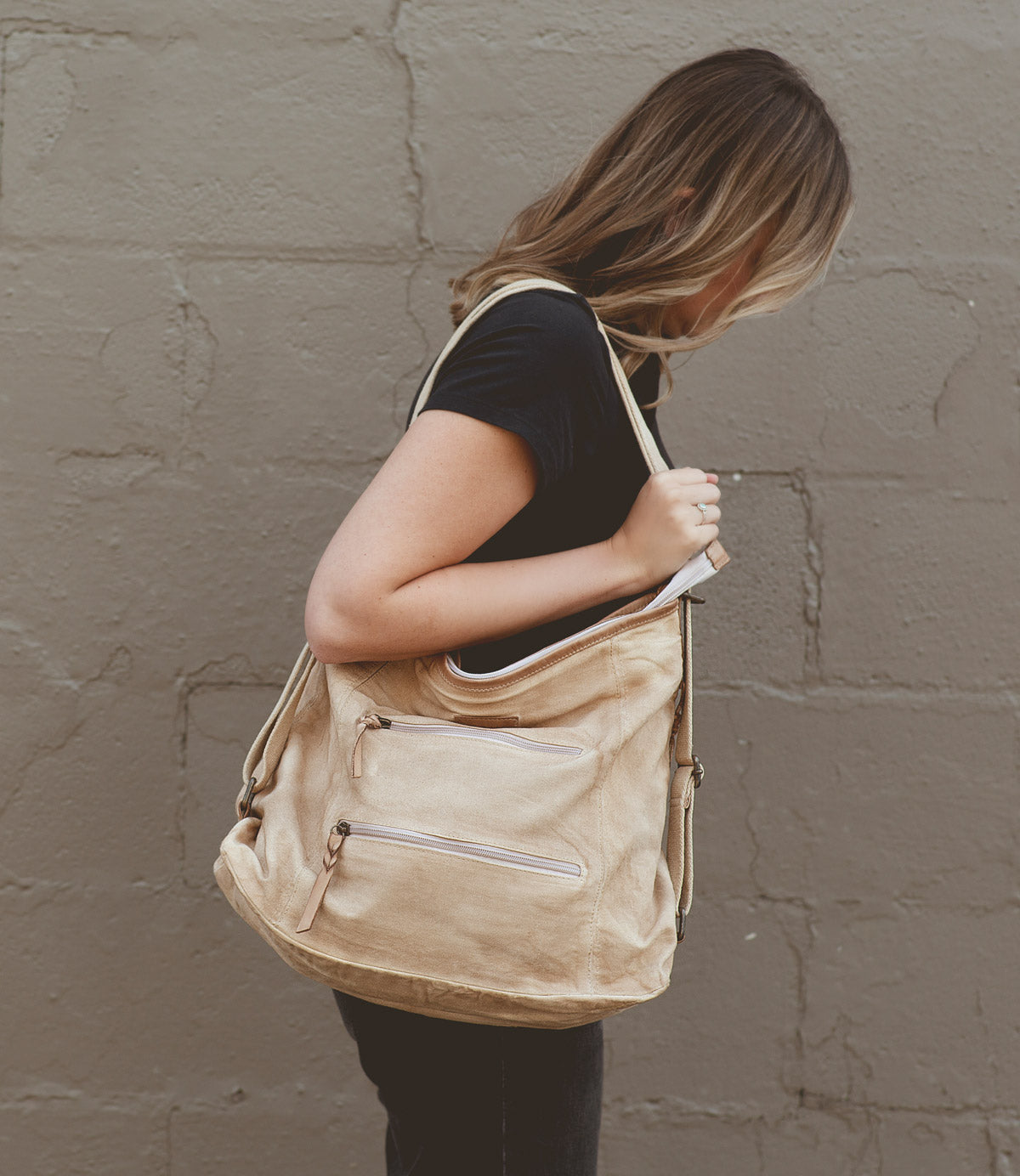 A person with long hair, dressed in a black shirt, stands sideways holding the Oganness by Roan, a large versatile bag with multiple zippers—an ideal travel companion—against a light textured wall.