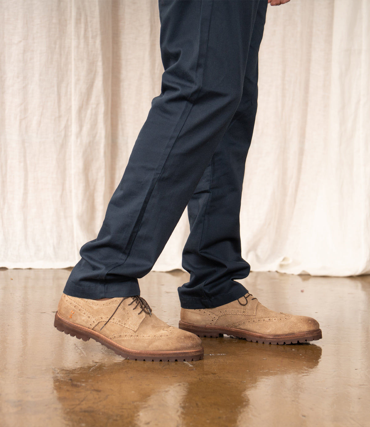 A person wearing navy blue pants and Roan Phiz tan wingtip brogue derby dress shoes stands on a polished brown floor with a light fabric backdrop.