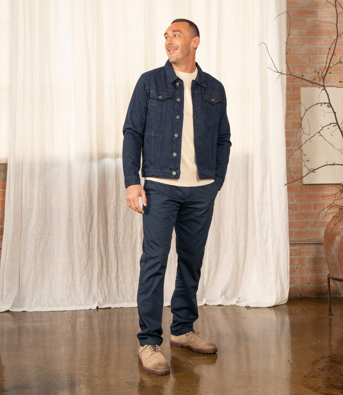 
                  
                    A man stands indoors on a polished floor, smiling to the side. He wears Roan's Phiz men's dress shoes with a denim jacket, beige shirt, and navy pants. White curtains and brick walls are in the background.
                  
                