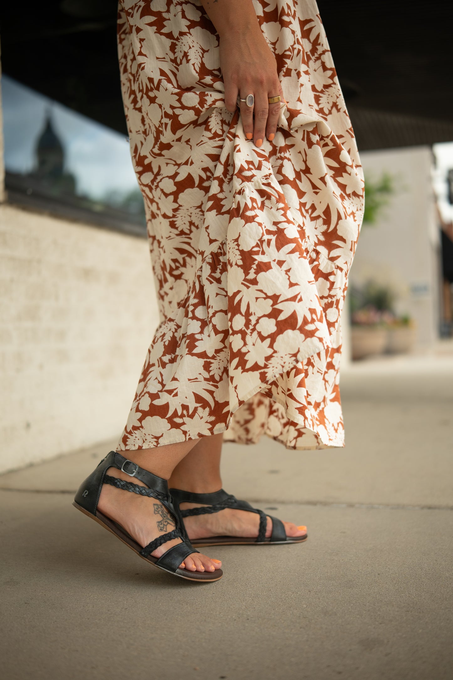 
                  
                    A person in a brown and white floral dress holds the hem, wearing Roan’s Posey black leather sandals with cushioned footbed, standing on a sidewalk.
                  
                