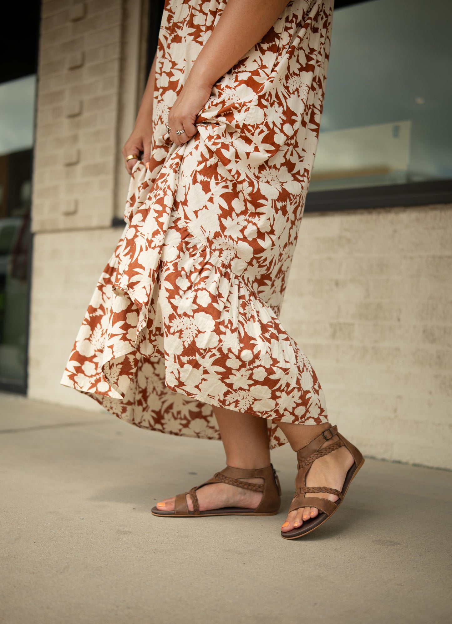 A person wearing the Posey brown and white floral dress by Roan and leather sandals with a cushioned footbed stands on a sidewalk near a light-colored brick wall.