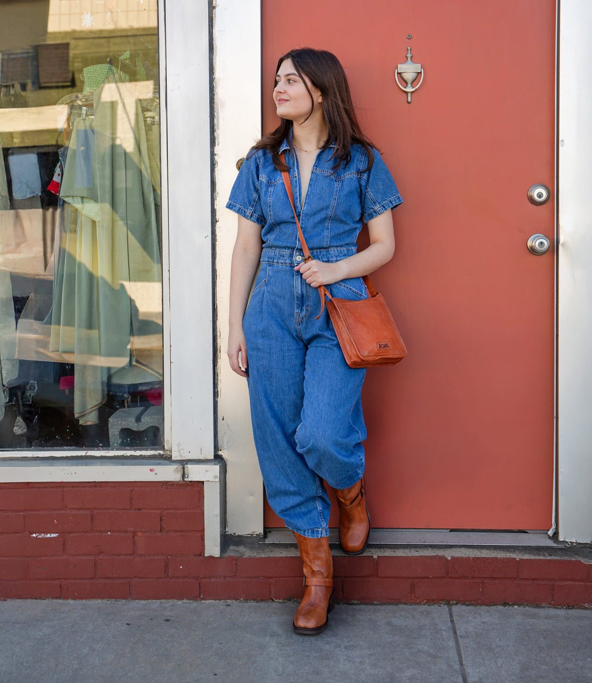 
                  
                    A woman in a denim jumpsuit and brown boots stands in front of a red door, carrying the Roan Quetzal bag and looking to the side.
                  
                