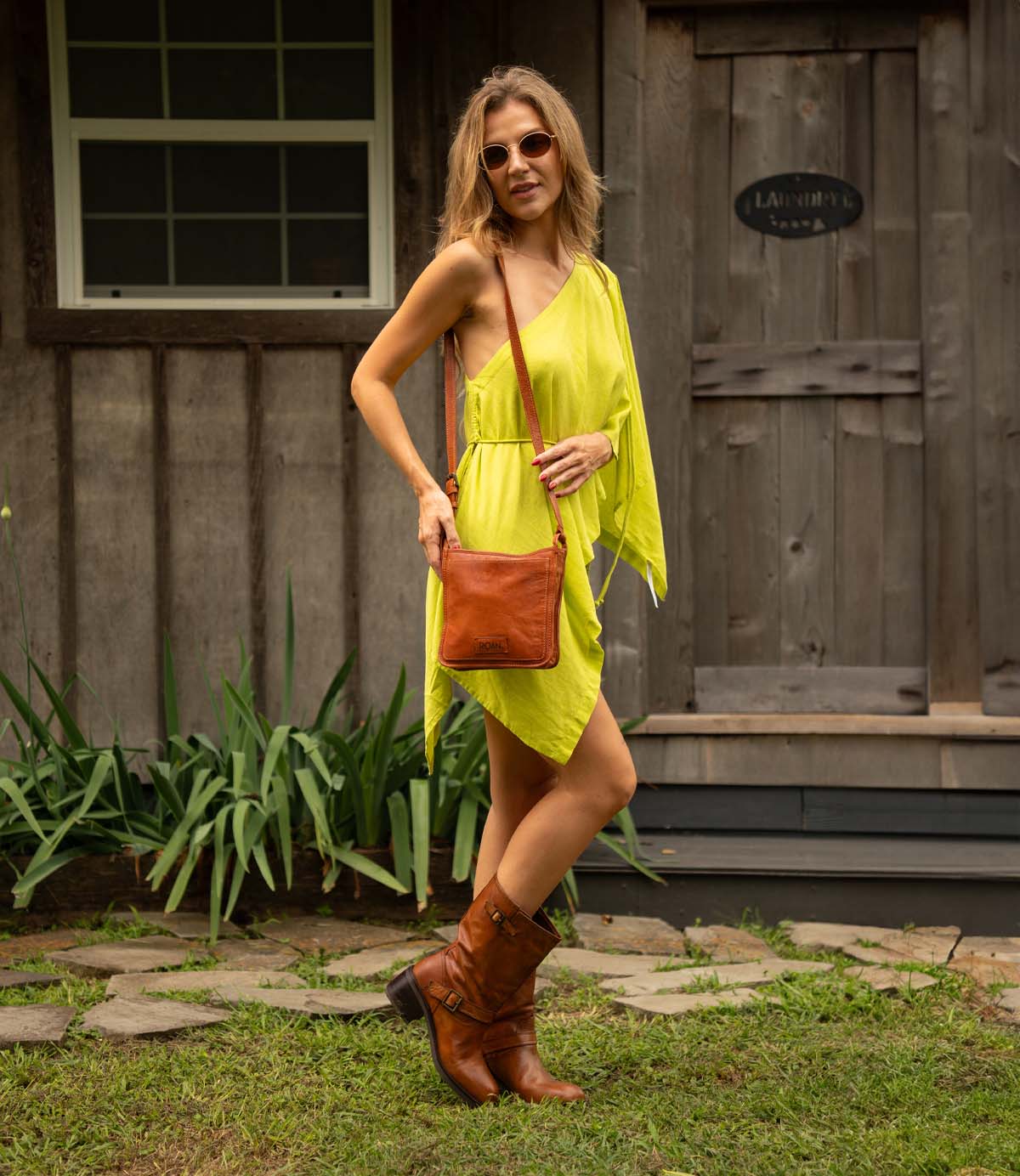 
                  
                    A woman in a yellow one-shoulder dress, brown boots, and sunglasses stands outside a wooden building, carrying the Roan Quetzal durable leather bag with a zip closure over her shoulder.
                  
                