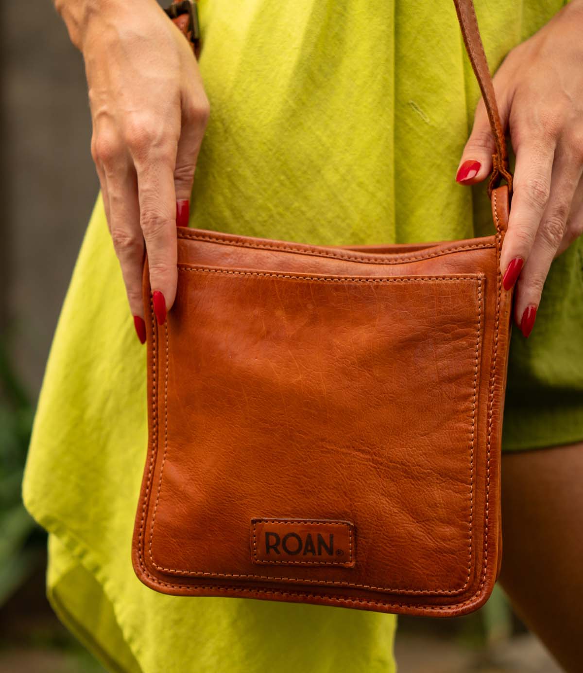 A person with red nail polish holds the durable leather Roan Quetzal crossbody bag with a zip closure, styled with a bright green dress—ideal for everyday use.