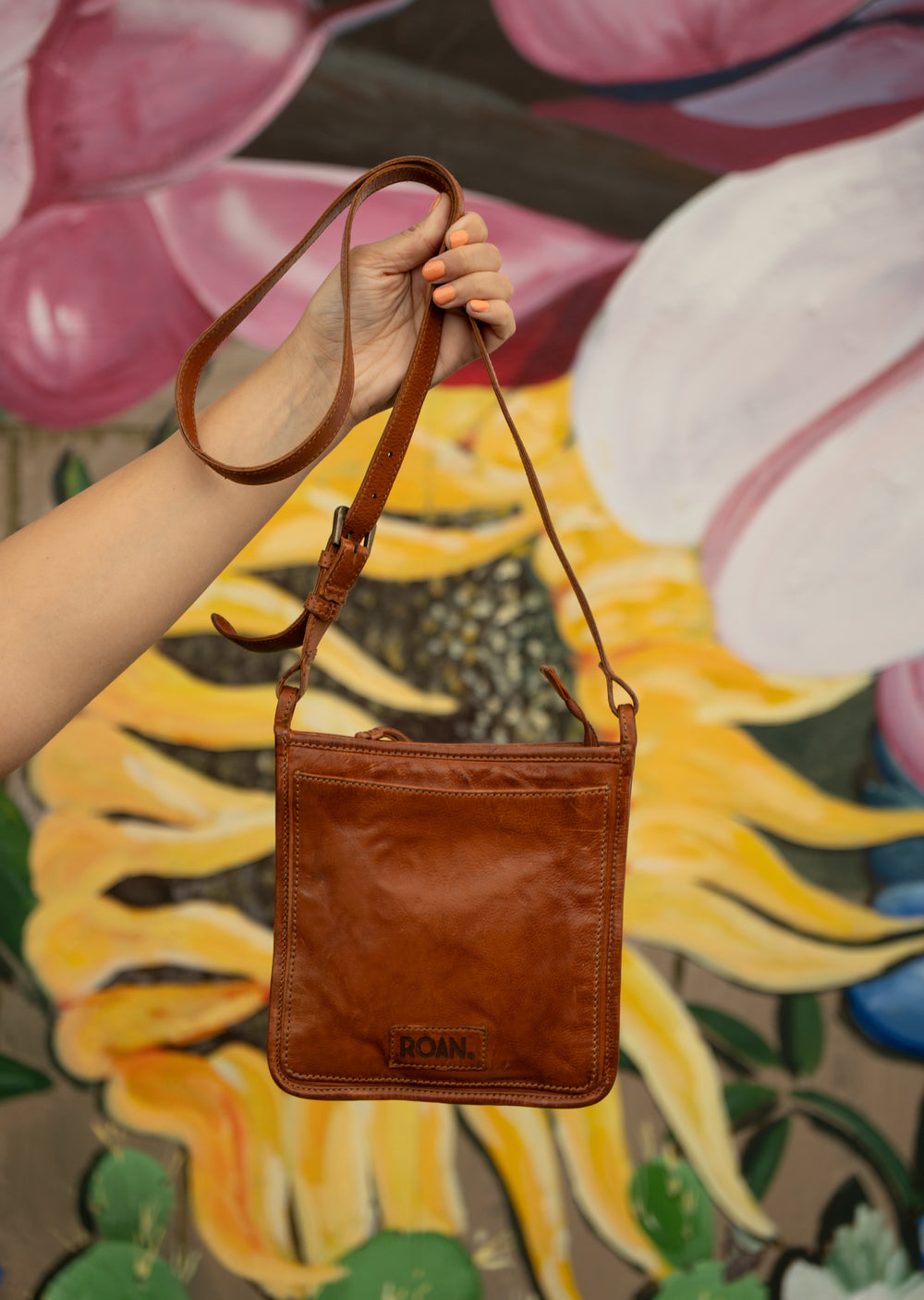 A hand holds up the Roan Quetzal bag, handcrafted from brown cow flame leather with a shoulder strap, in front of a colorful floral mural.