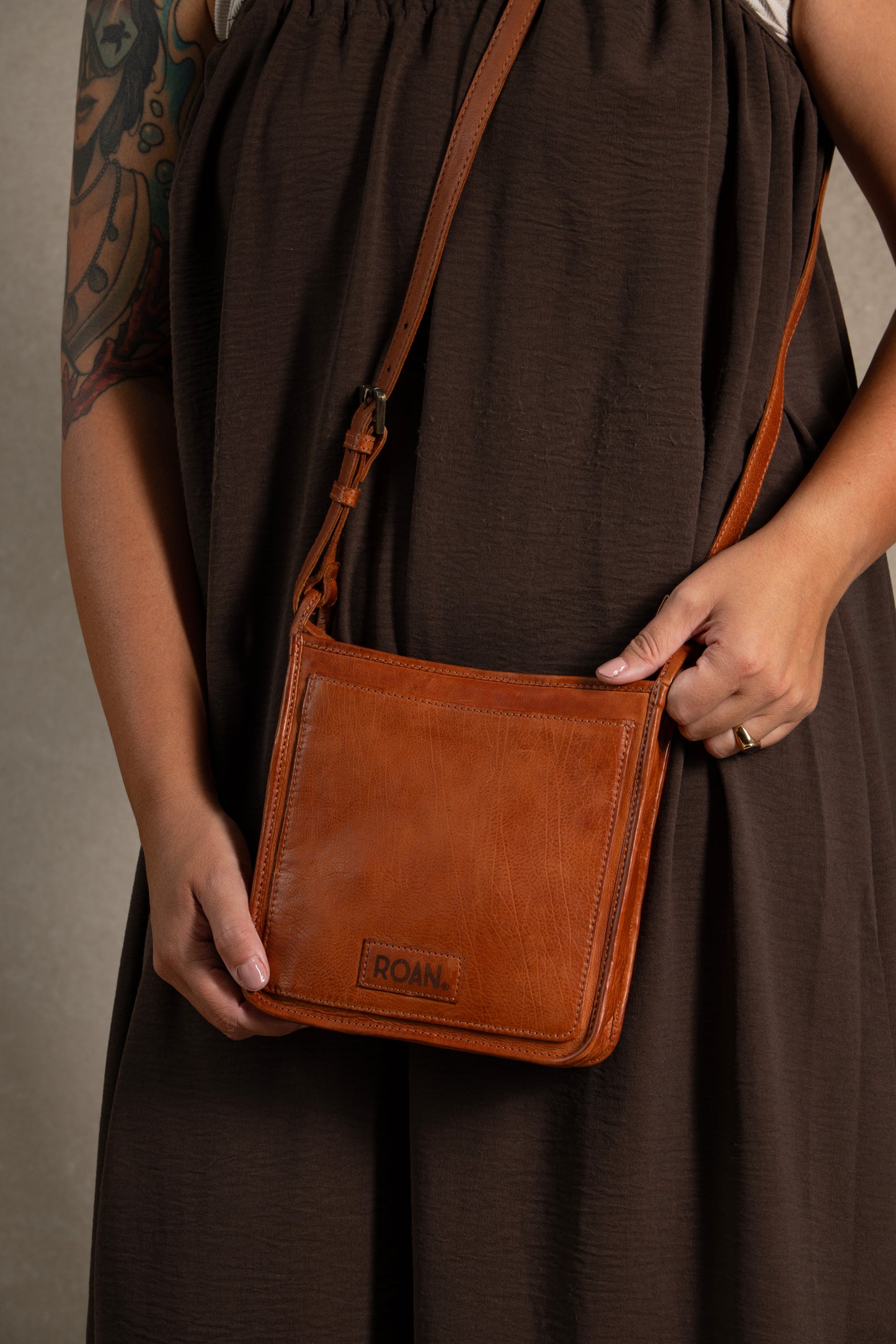 A person in a brown dress holds the Quetzal—a durable leather bag with a zip closure and “Roan” elegantly embossed on the front—perfect for everyday use.