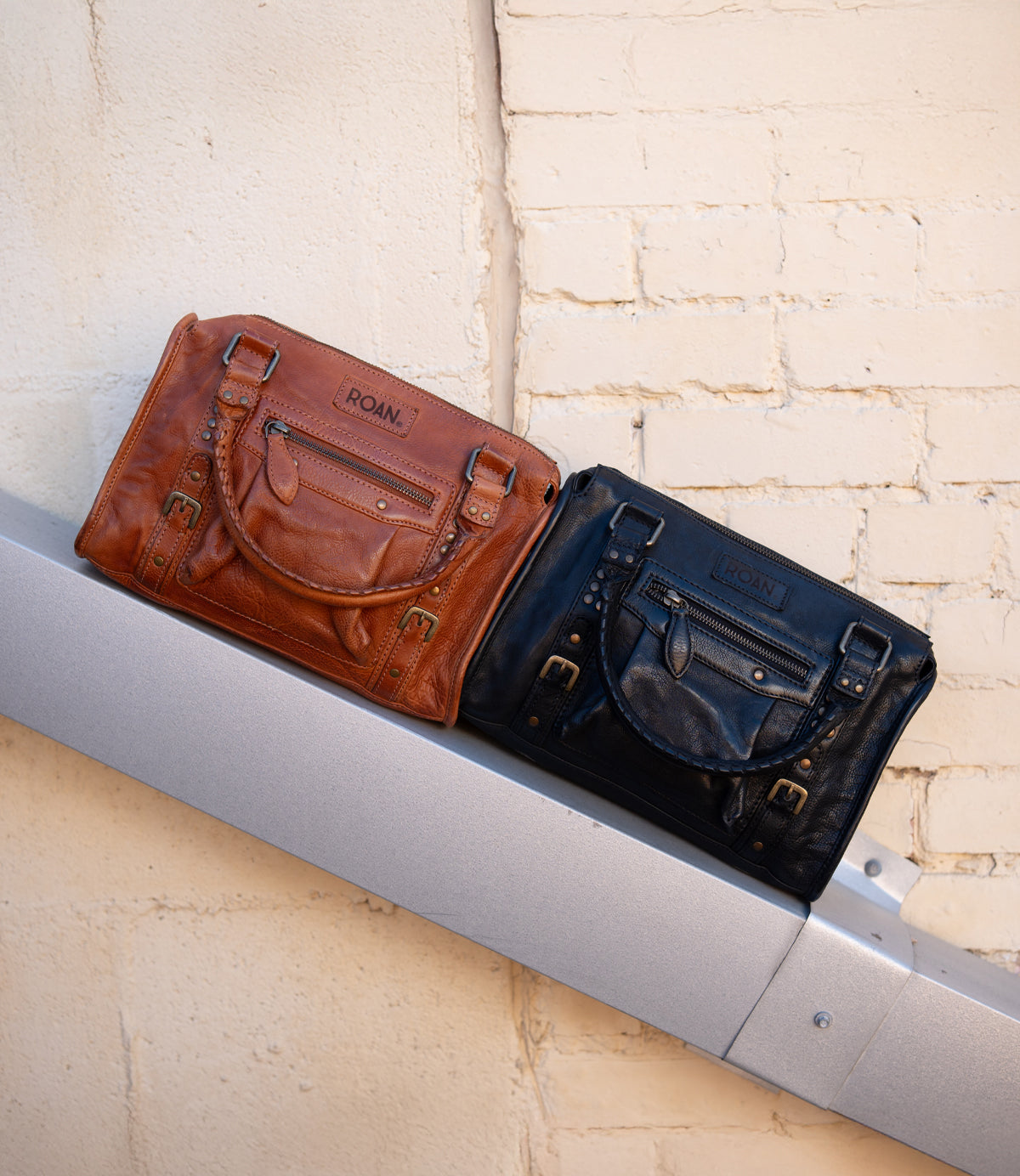 
                  
                    Two Roan Visual leather satchels—one brown, one black with subtle studs—are displayed side by side on a metal beam against a beige brick wall.
                  
                