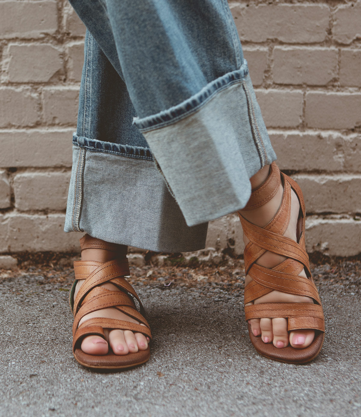 Close-up of a person wearing wide-cuff denim pants and Roan Royalty brown leather sandals with a cushioned footbed, standing on concrete ground against a brick wall background.