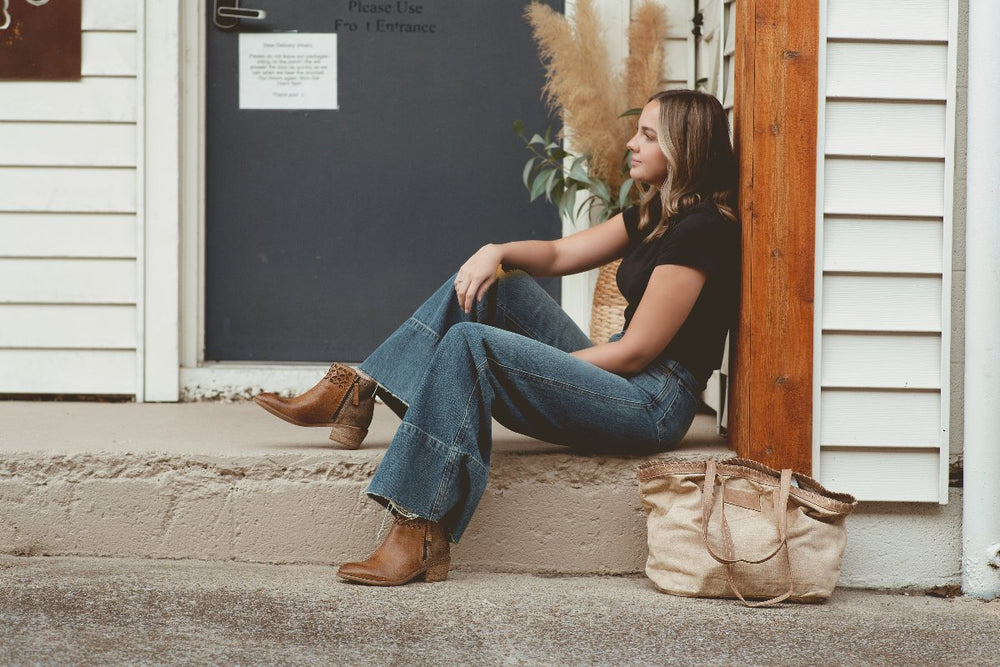 A woman in jeans and a black shirt sits on a step next to a door, with the Roan Rutherford handmade tote placed on the ground beside her, perfectly complementing her versatile lifestyle.