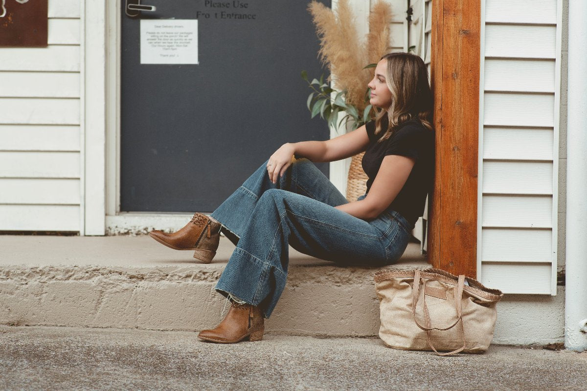 A woman in jeans and a black shirt sits on a step next to a door, with the Roan Rutherford handmade tote placed on the ground beside her, perfectly complementing her versatile lifestyle.