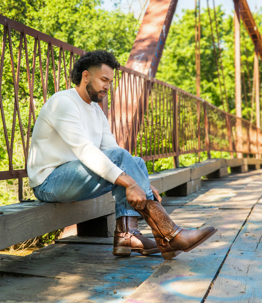 A man sits on a wooden bridge, wearing jeans and a white sweater as he puts on Roan's Saber boots—brown, full-grain leather with a square toe. Trees and metal railings are in the background.