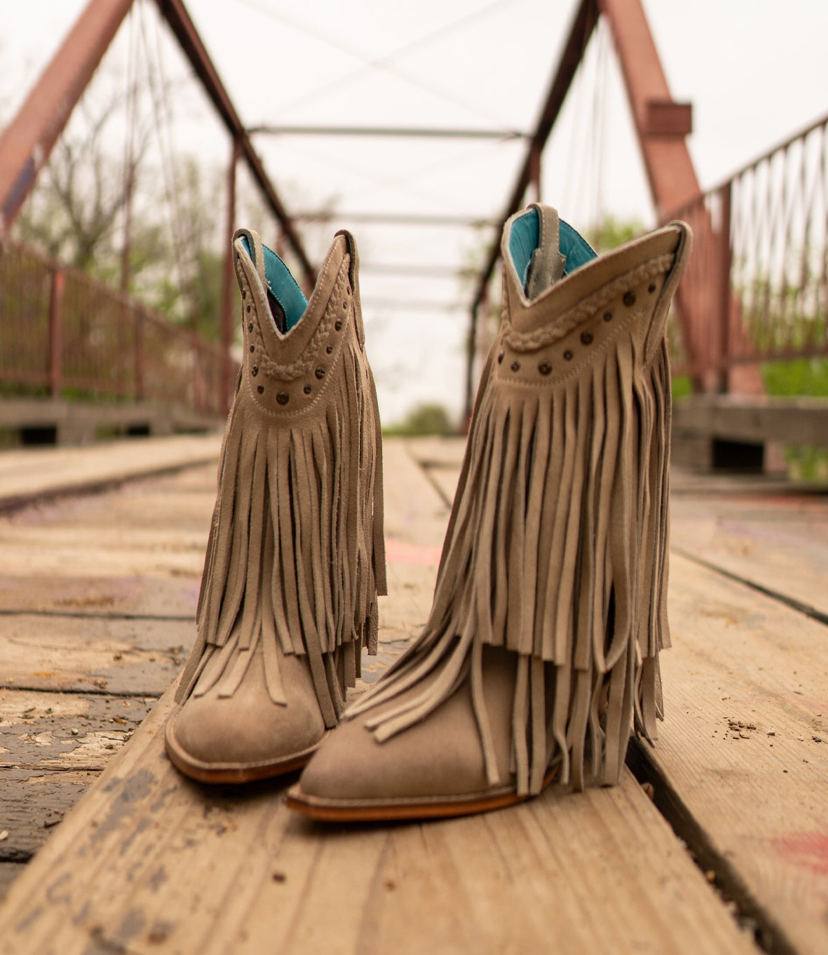 Suede cowboy boots named "Setup" by Roan, stand on a wooden bridge with metal railings and trusses in the background, exuding a Western-inspired flair.