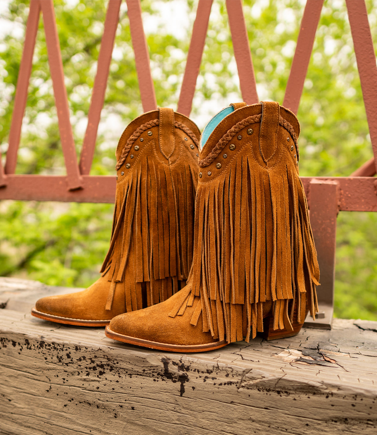 
                  
                    Roan's "Setup" suede boots, featuring fringe and stud details, are displayed on a wooden surface against a pink metal fence with lush green foliage.
                  
                