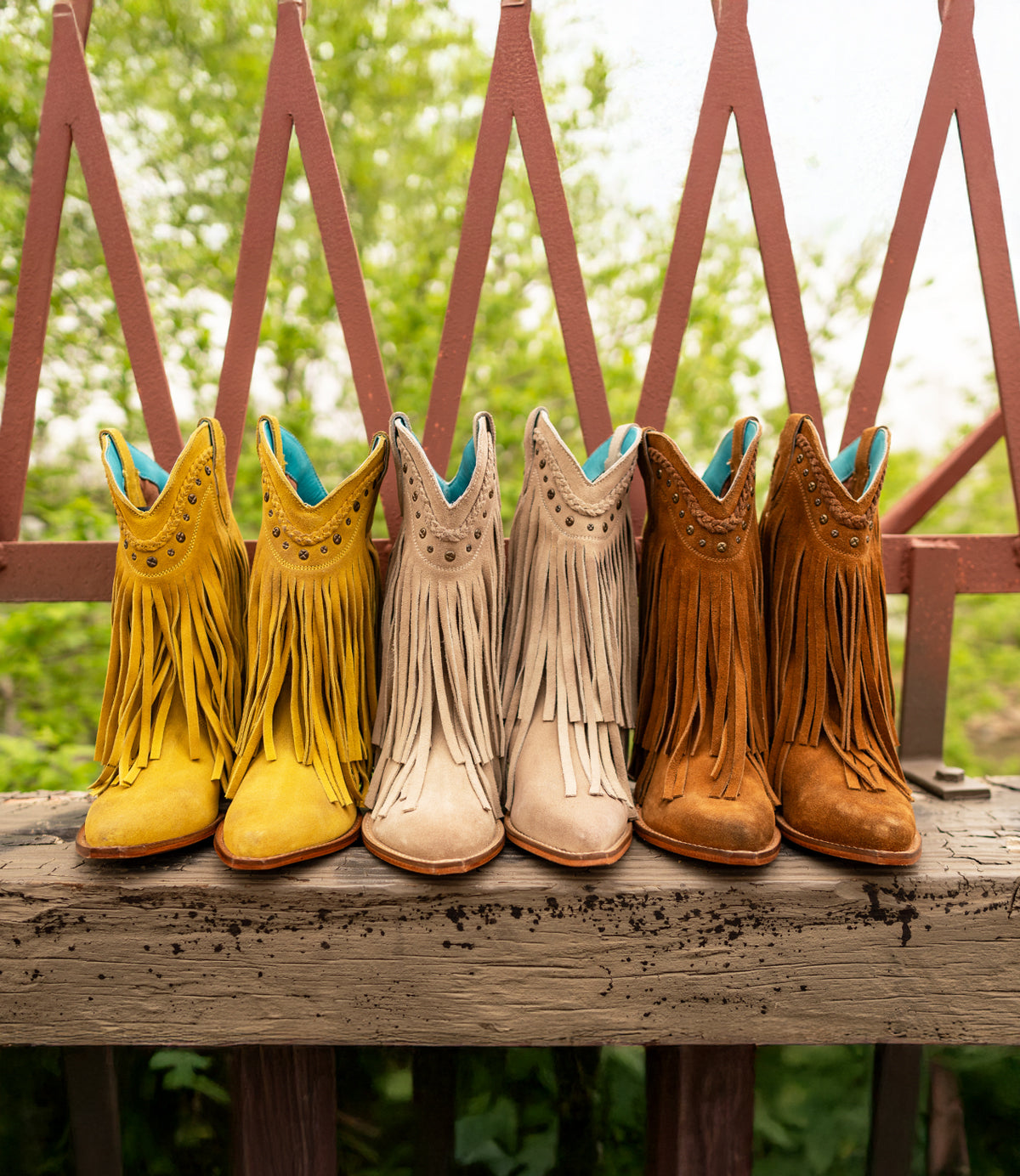 
                  
                    Three pairs of Roan's 'Setup' boots in yellow, beige, and brown sit on a wooden surface. Their fringe detailing adds charm against greenery and a pink fence backdrop.
                  
                