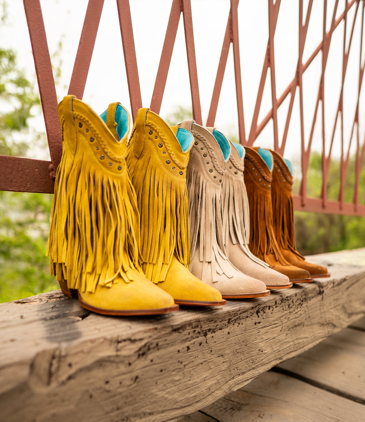 Four pairs of Roan's Setup boots, featuring Western-inspired designs with fringe detailing in yellow, beige, and brown, are lined up on a wooden beam near a metal railing.