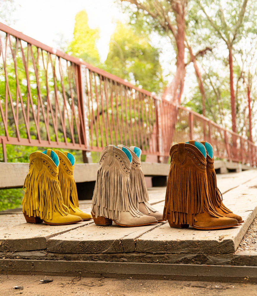 
                  
                    Three pairs of Roan's Western-inspired Setup boots in yellow, beige, and brown are arranged on a wooden bridge. Each suede boot showcases eye-catching fringe detailing, framed by metal railings and softly swaying trees in the background.
                  
                