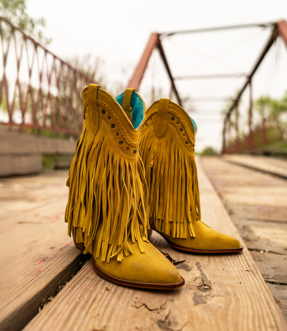 
                  
                    Roan's Setup cowboy boots in yellow suede with fringe details are showcased on a wooden bridge featuring a rust-colored metal railing.
                  
                