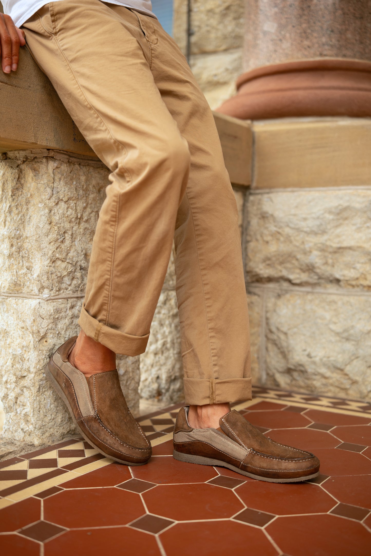 
                  
                    A person in light brown pants wears Roan’s Shevon brown leather slip-on shoes with a cushioned insole, leaning against a stone wall on patterned tile flooring.
                  
                