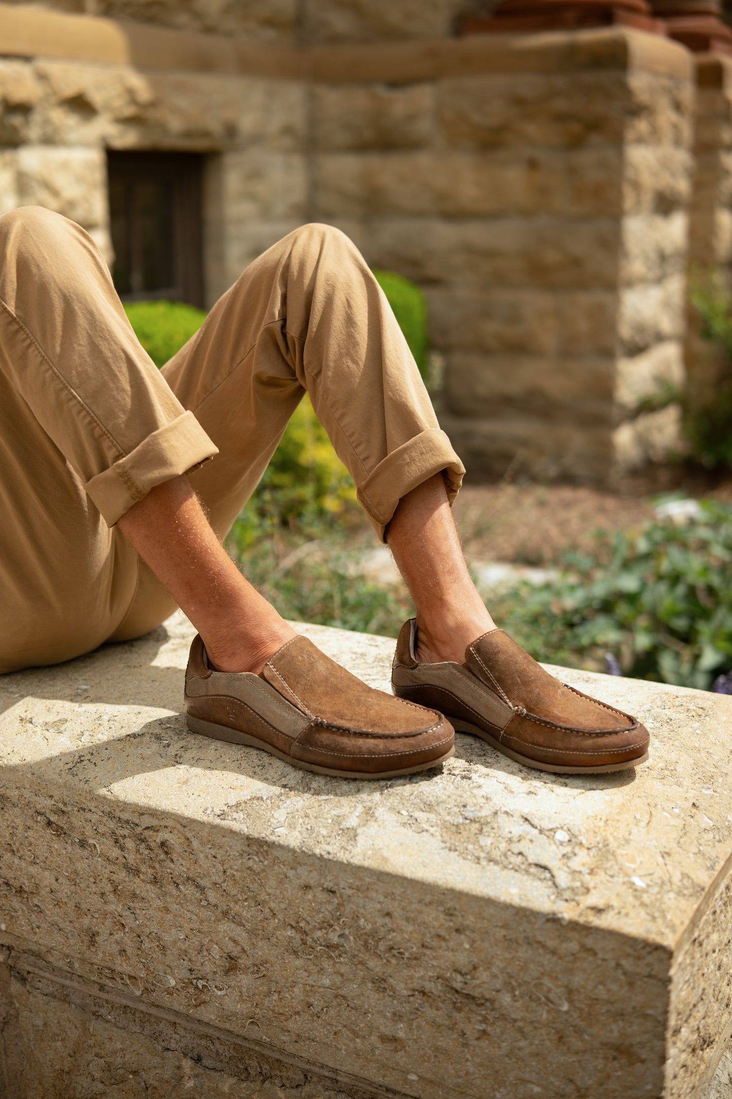 
                  
                    A person sits outdoors on a stone ledge, legs crossed at the ankles, wearing tan pants and Roan’s Shevon brown leather slip-on shoes featuring a cushioned insole.
                  
                