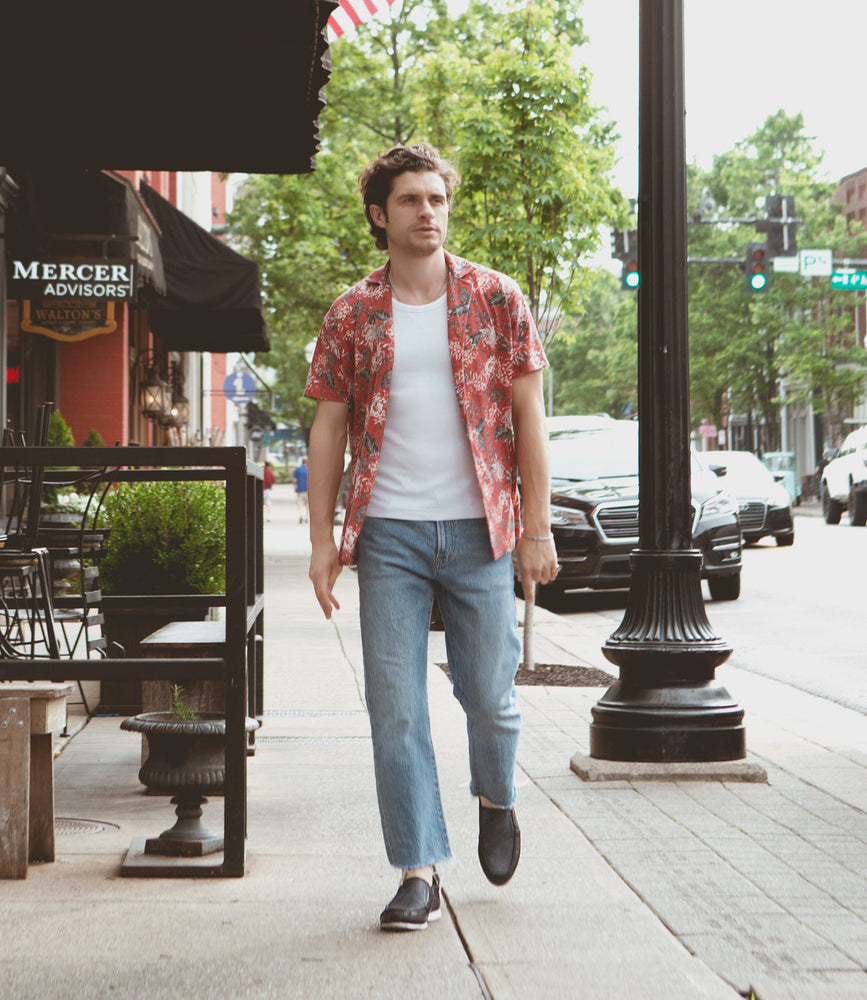 
                  
                    A man in a floral print shirt and jeans, sporting Roan Shevon leather slip-ons with cushioned insoles, walks on a city sidewalk near parked cars and buildings.
                  
                