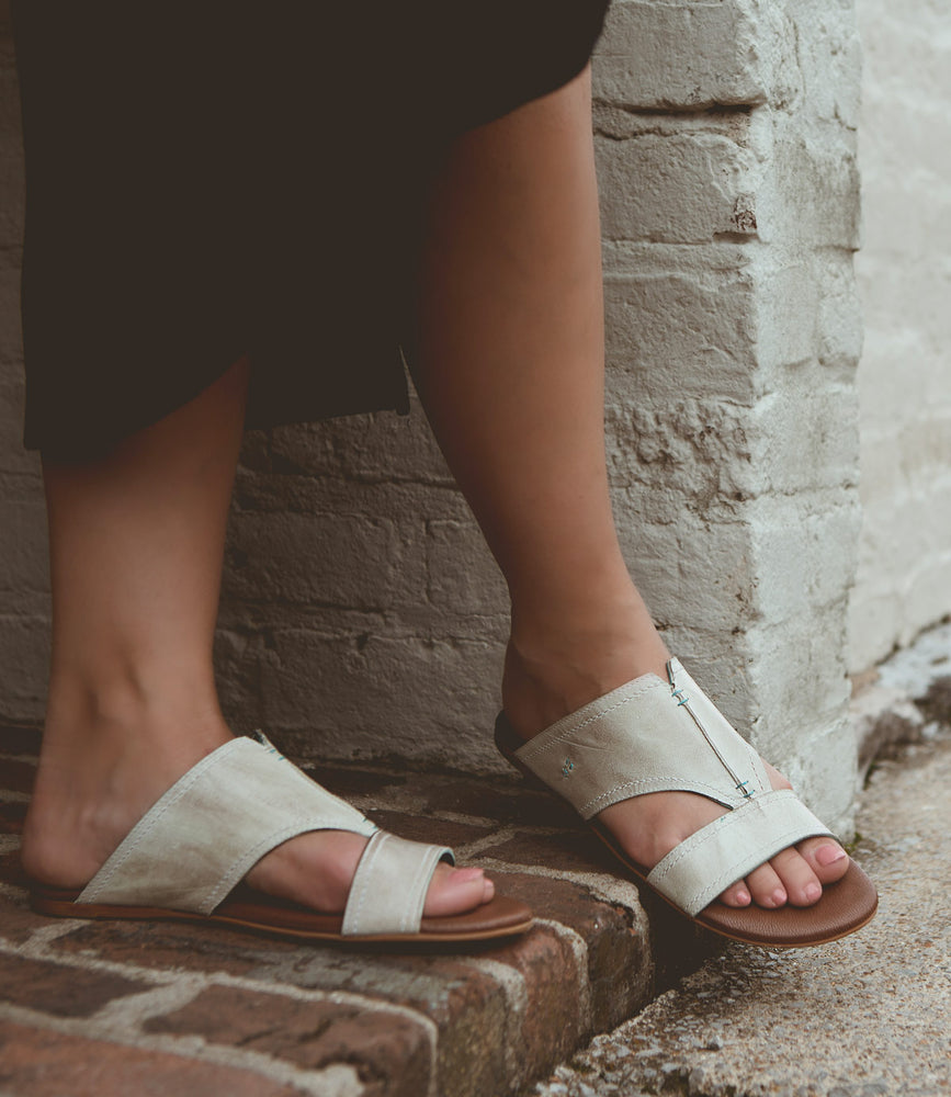 Person wearing all-leather, light-colored Roan Somerville summer slide sandals with a split toe design, standing on a brick surface near a white brick wall.