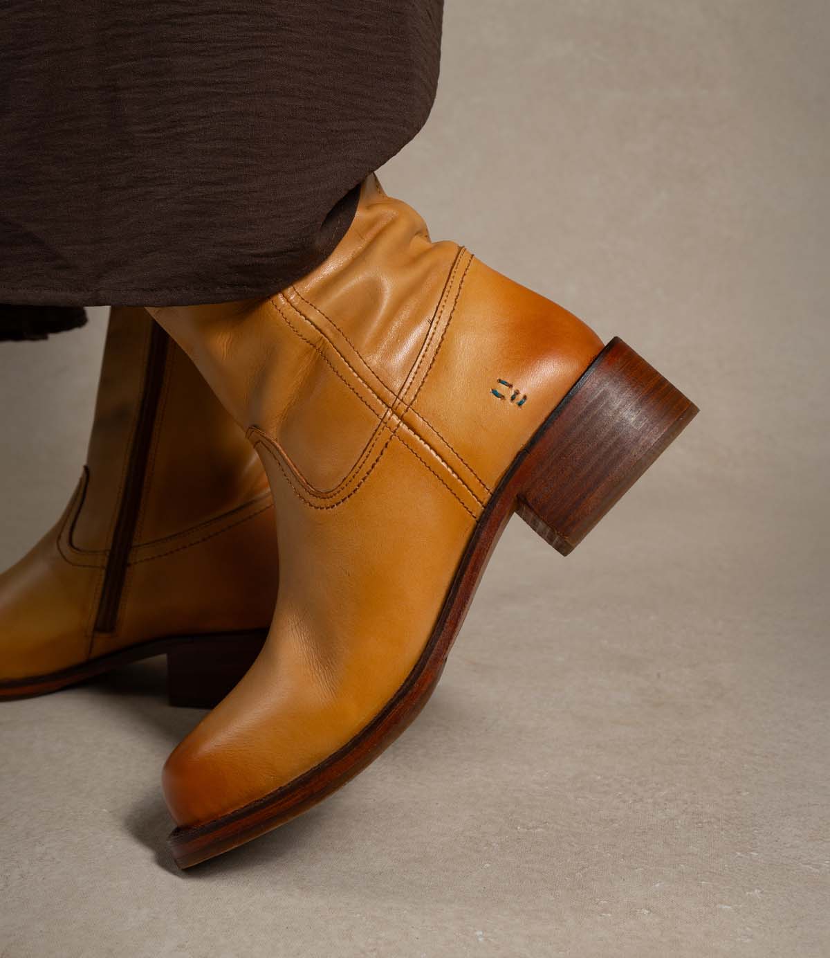 
                  
                    A person wearing the Roan Splitter tan leather ankle boots, which feature a chunky leather silhouette, a wooden heel, and an inner-ankle zipper, stands on a light beige surface in dark brown pants.
                  
                