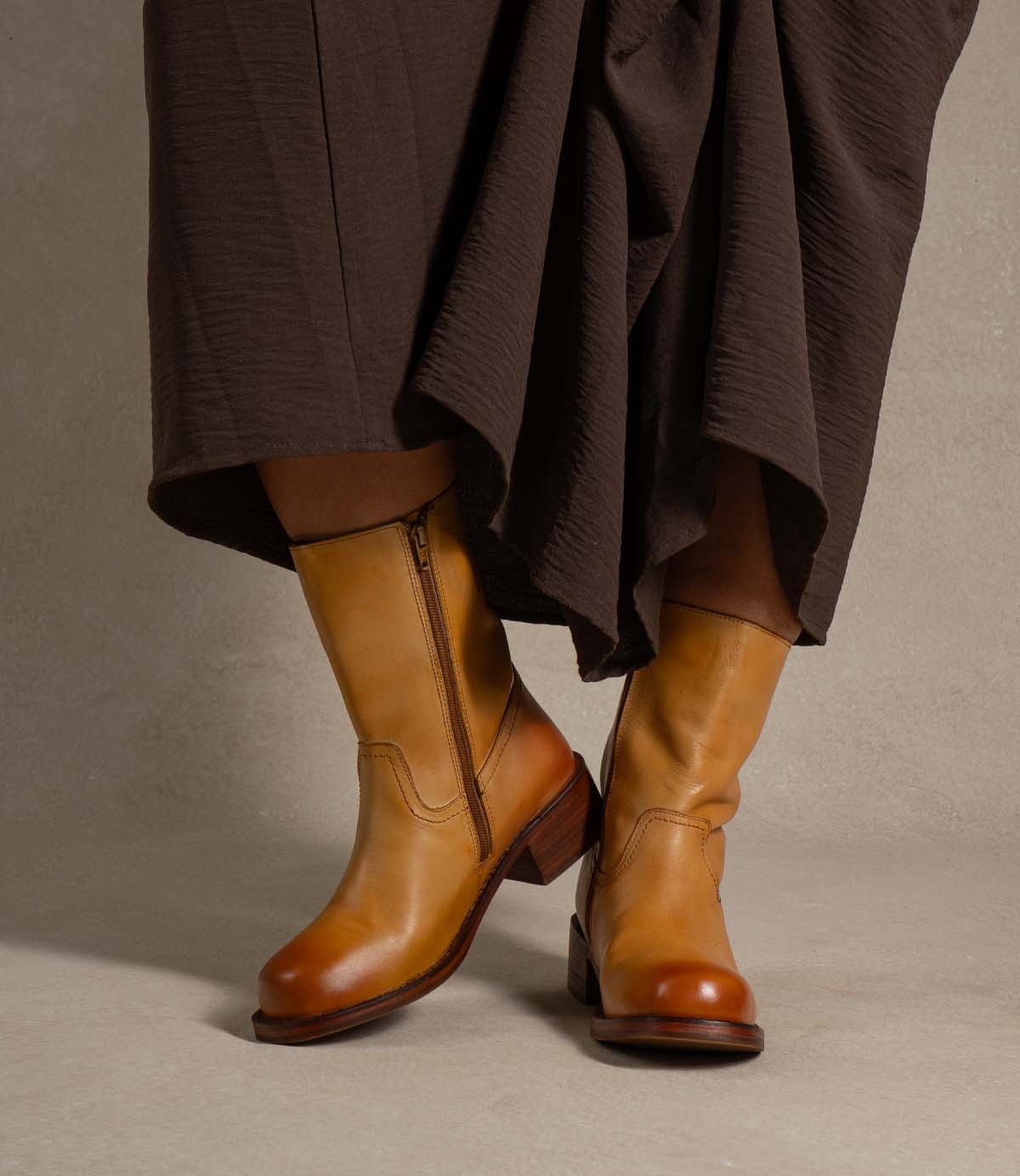 
                  
                    A person wearing a brown skirt and Roan Splitter tan leather mid-calf boots, featuring a sturdy, chunky leather silhouette and an inner-ankle zipper, stands on a neutral surface.
                  
                