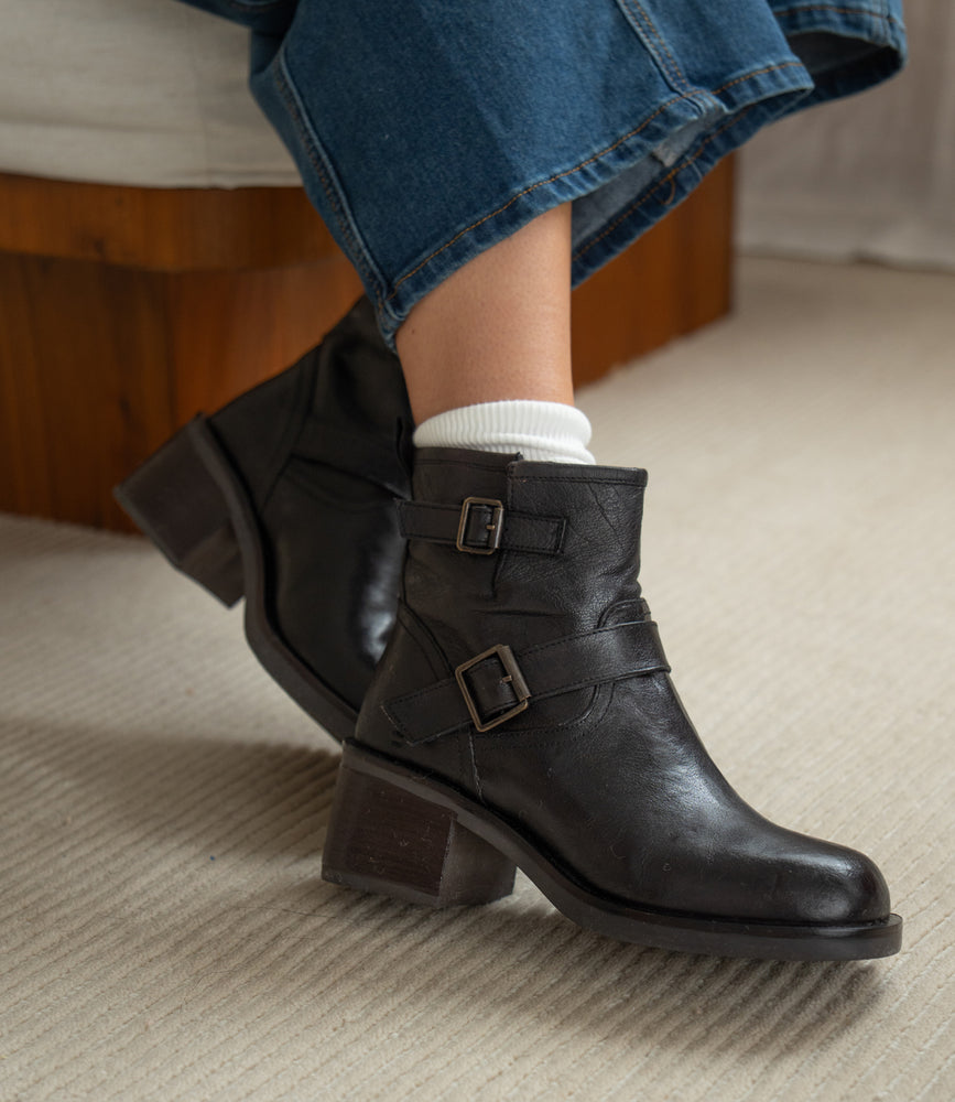 Wearing white socks and Spry moto-inspired ankle boots by Roan—featuring double buckle straps, block heels, and hand-finished leather—a person in blue jeans sits on a carpeted floor.