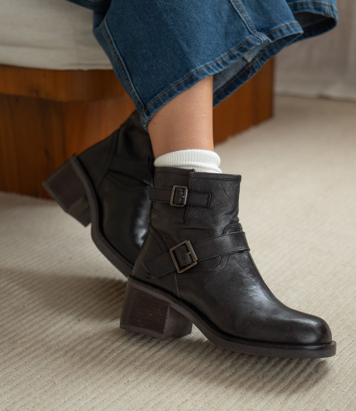Wearing white socks and Spry moto-inspired ankle boots by Roan—featuring double buckle straps, block heels, and hand-finished leather—a person in blue jeans sits on a carpeted floor.