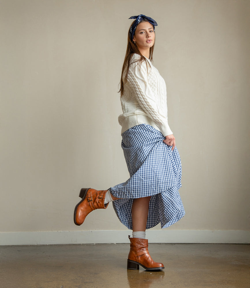 
                  
                    A woman wearing Roan’s Spry moto-inspired ankle boots in hand-finished leather stands on one leg against a beige wall, dressed in a cream sweater, blue gingham skirt, and blue headband.
                  
                