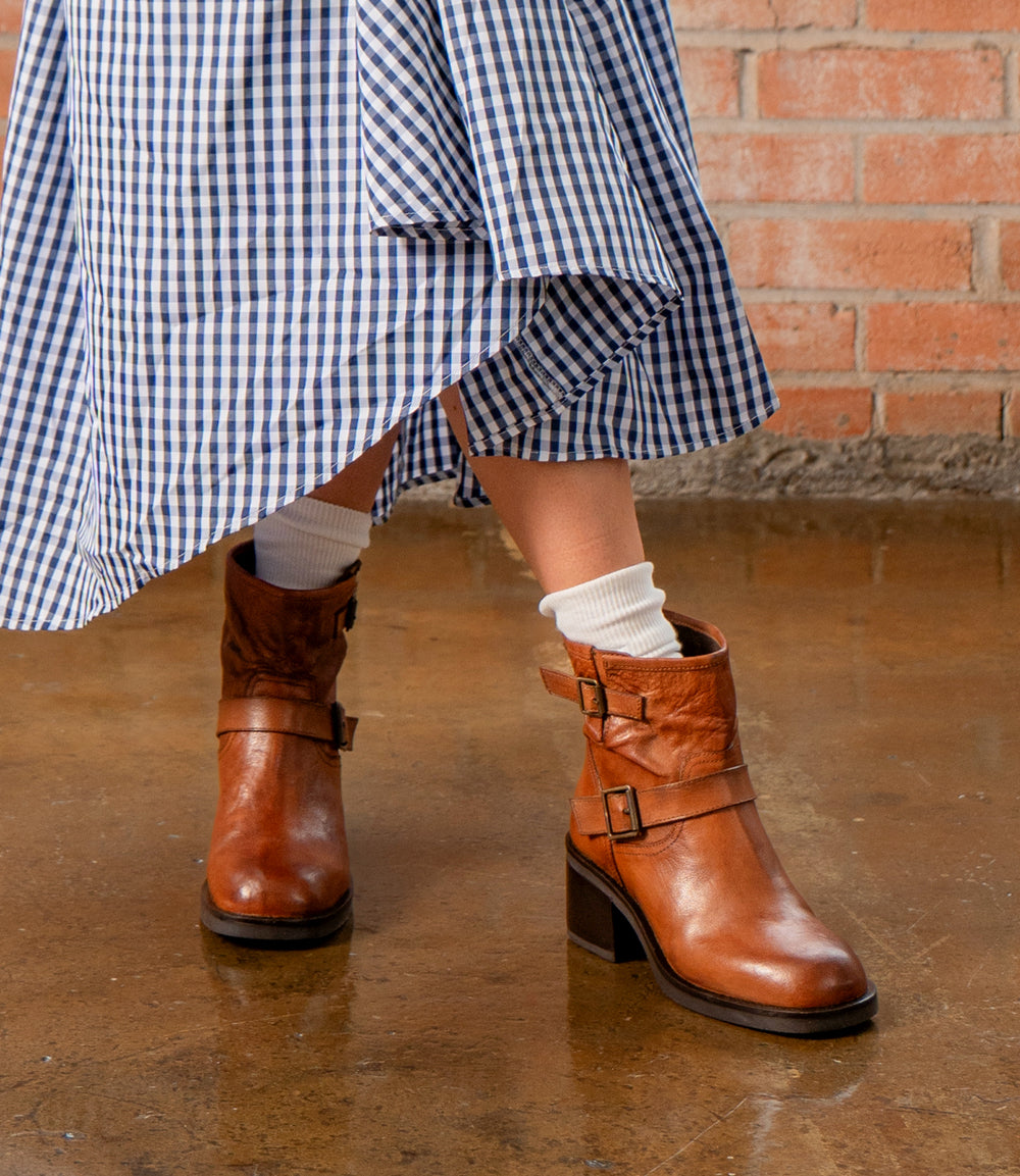A person wearing the Spry moto-inspired ankle boots with a double buckle strap by Roan stands on a polished floor in front of a brick wall, styled with a blue and white gingham skirt and white socks.