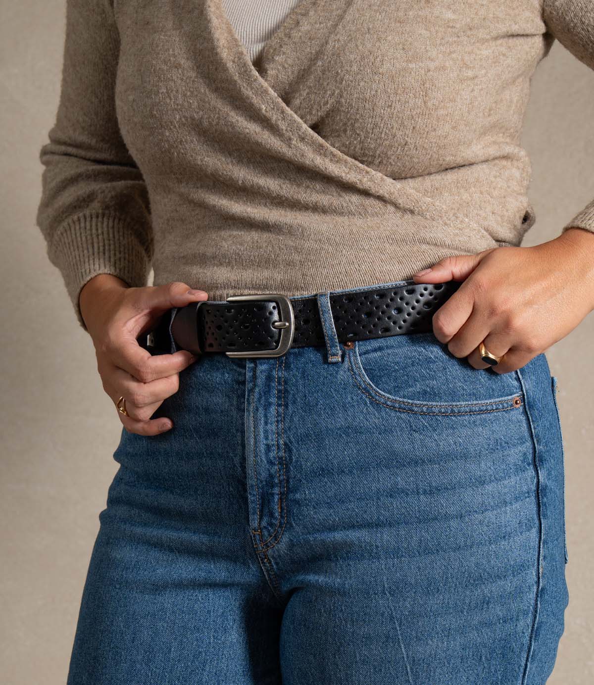 A person wearing a beige sweater and blue jeans adjusts the Roan Spurline black belt, featuring a perforated pattern and metal buckle, using both hands.