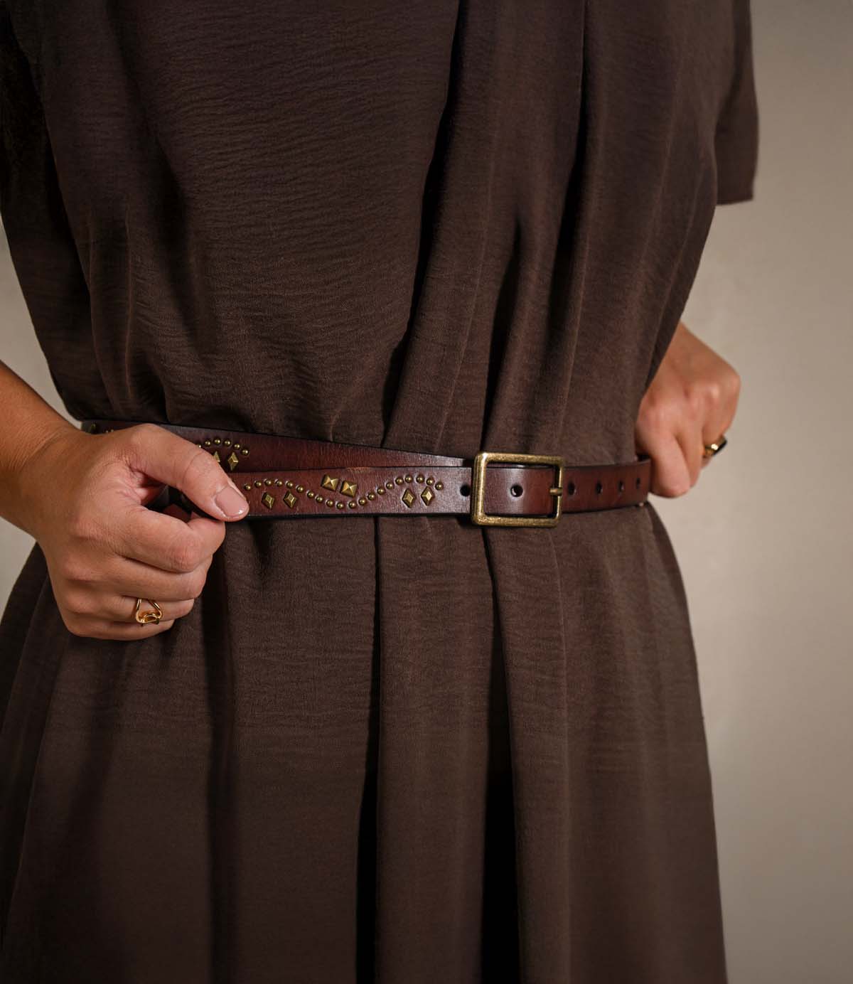 Wearing a loose brown dress, the person fastens the Roan Tackson studded leather belt with decorative metalwork and a rectangular buckle around their waist.