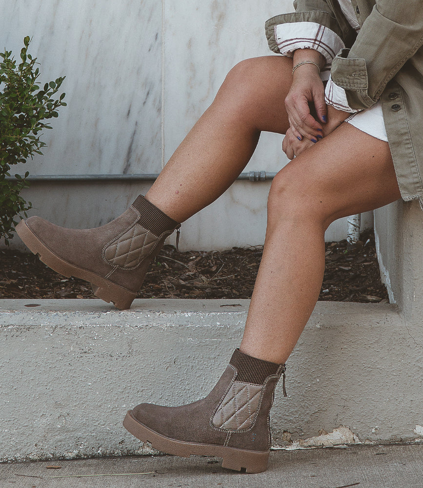 Seated on concrete steps surrounded by lush greenery, a person exudes versatile style by pairing Roan's "Thoughtful" brown boots with a khaki coat and white dress.