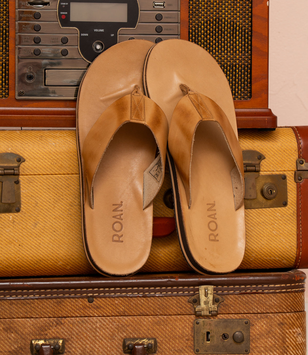 A pair of Townfolk flip-flops in brown, featuring "Roan" on the soles, exude rustic charm as they rest on a vintage suitcase and stereo system.