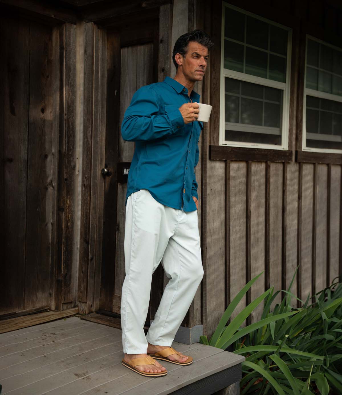 A man in a teal shirt, white pants, and Roan Townfolk leather thong sandals with a padded footbed stands on a wooden porch with green plants at his side and two windows behind him, holding a mug.