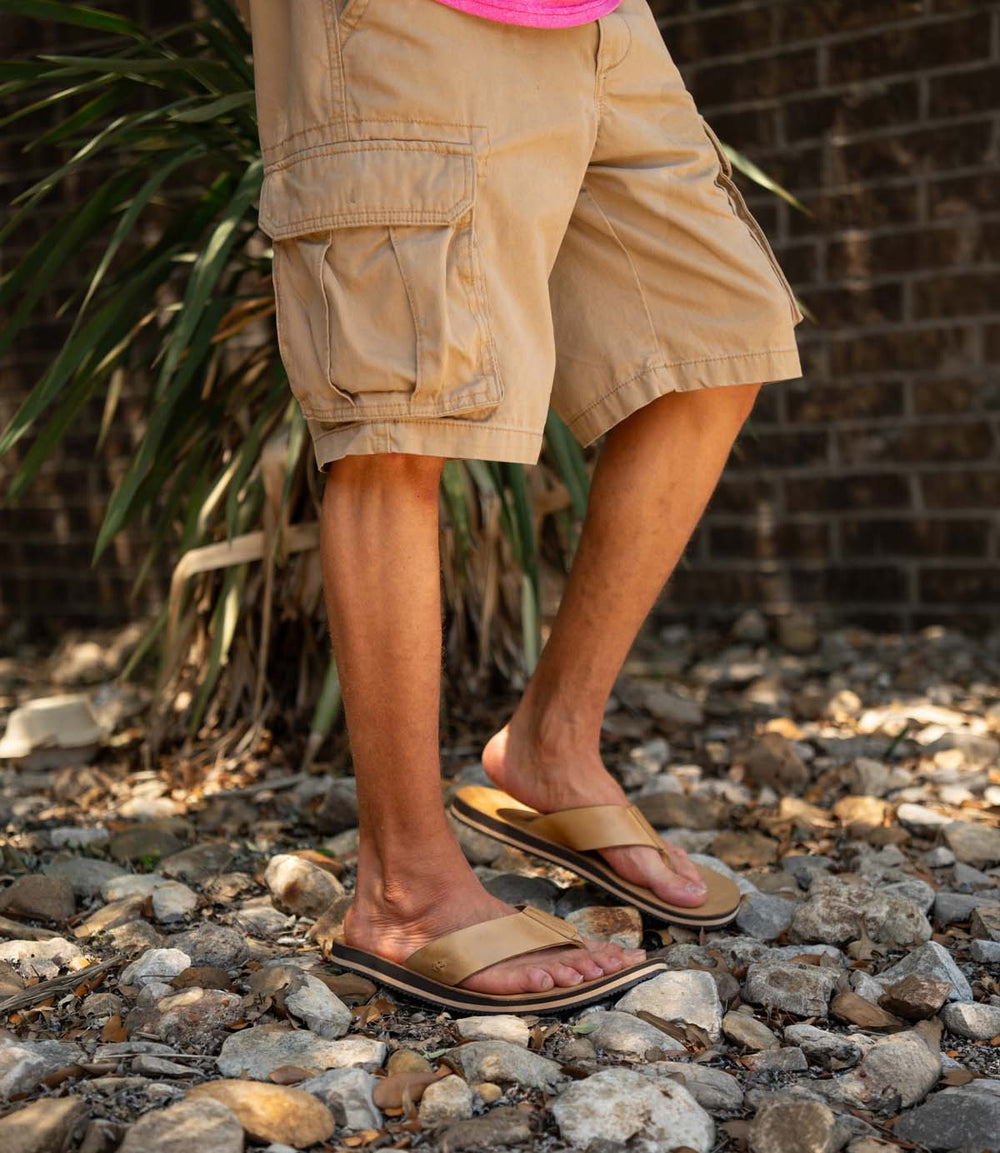 Wearing Roan's Townfolk full-grain leather sandals with a padded leather footbed and tan cargo shorts, this person stands on rocky ground amid plants and a brick wall.