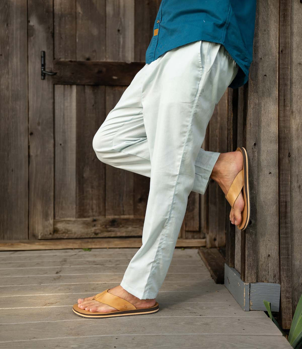 Wearing Roan's Townfolk tan leather thong sandals with padded footbed, a person in light pants and a teal shirt leans against a wooden wall on a porch with a closed wooden door behind them.