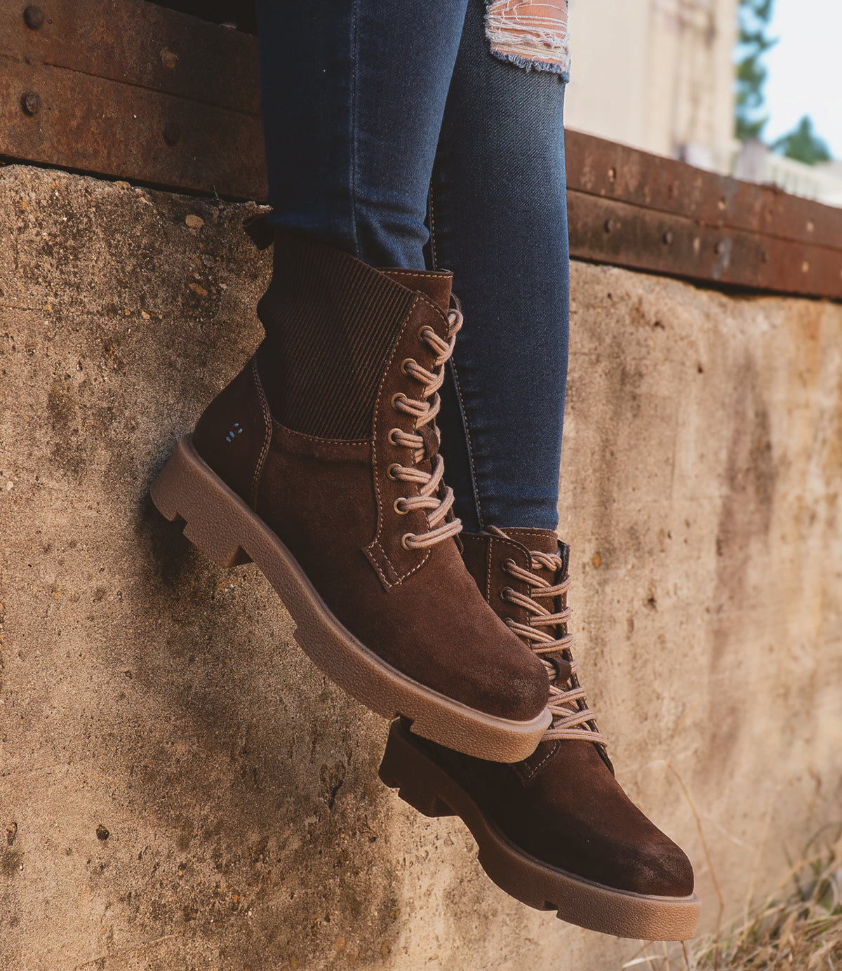A person wearing Roan's Transform brown lace-up boots with a flannel lining and blue jeans is sitting on a concrete ledge.
