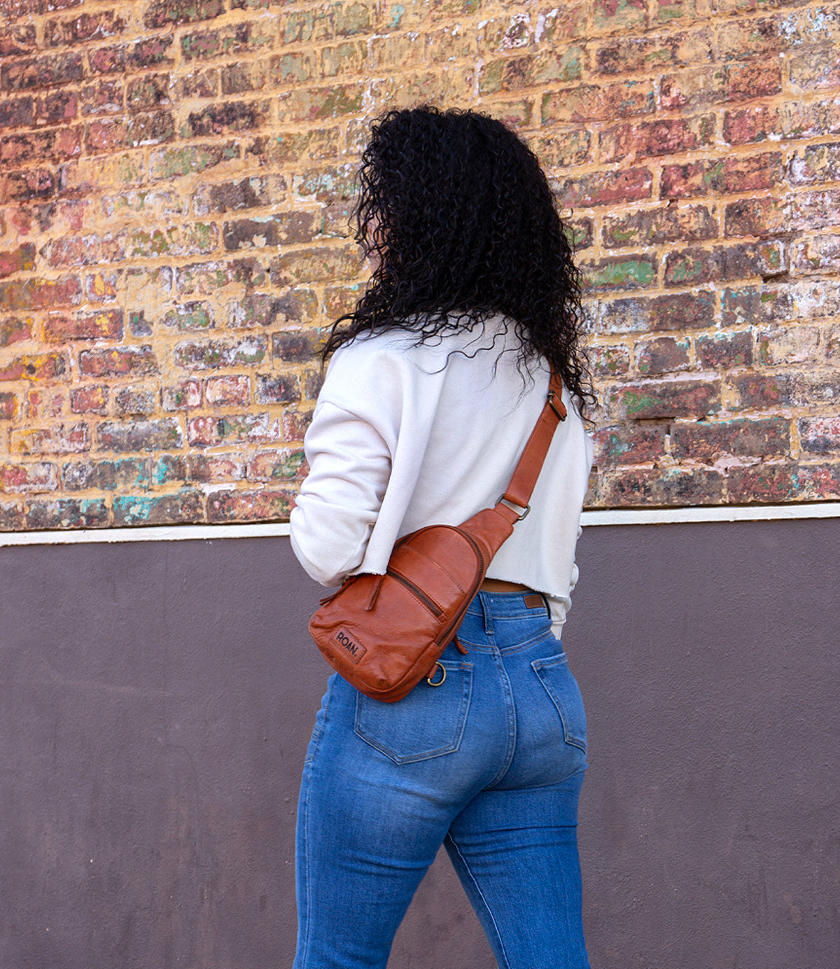 
                  
                    A person with curly hair, in a light sweater and blue jeans, stands facing a brick wall, carrying Roan’s Trickle—a compact leather bag that adds versatile style to their everyday look.
                  
                