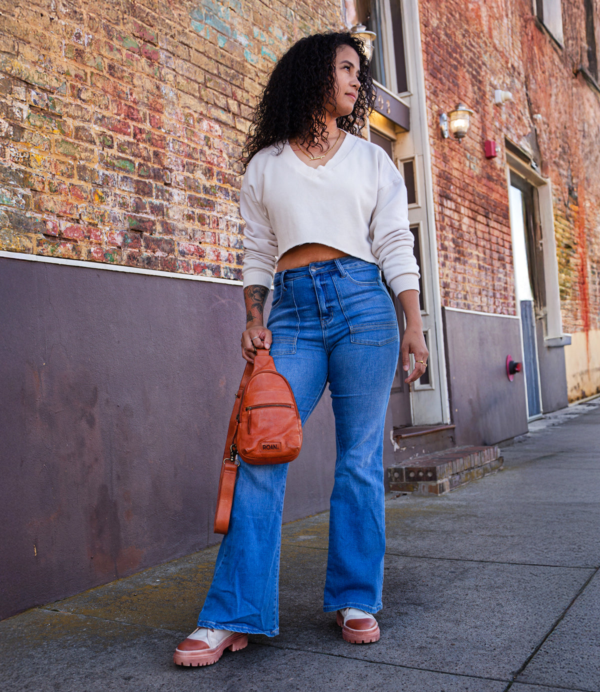 
                  
                    A person with curly hair stands on a sidewalk by a brick wall, wearing a white cropped sweater, blue jeans, pink sandals, and carrying the Roan Trickle bag for versatile everyday style.
                  
                