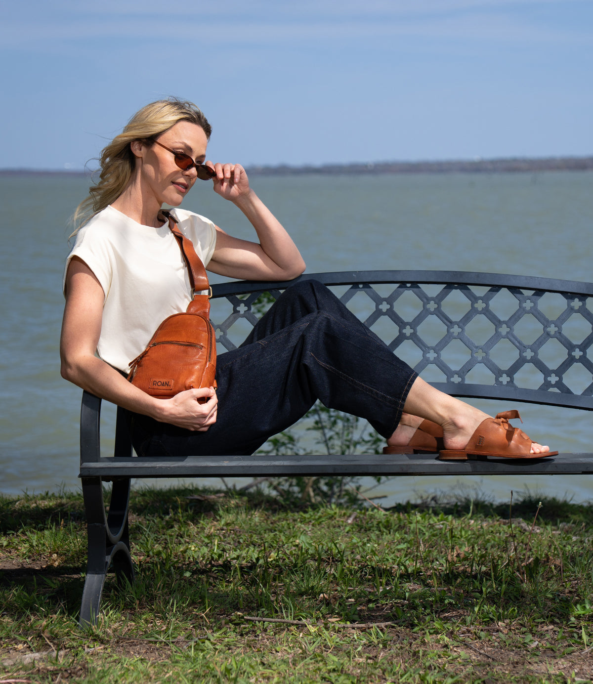 A woman sits on a metal bench by the water, wearing sunglasses, a white top, dark pants, brown sandals, and carrying Roan's Trickle—a compact leather bag that brings everyday versatility to her relaxed style.