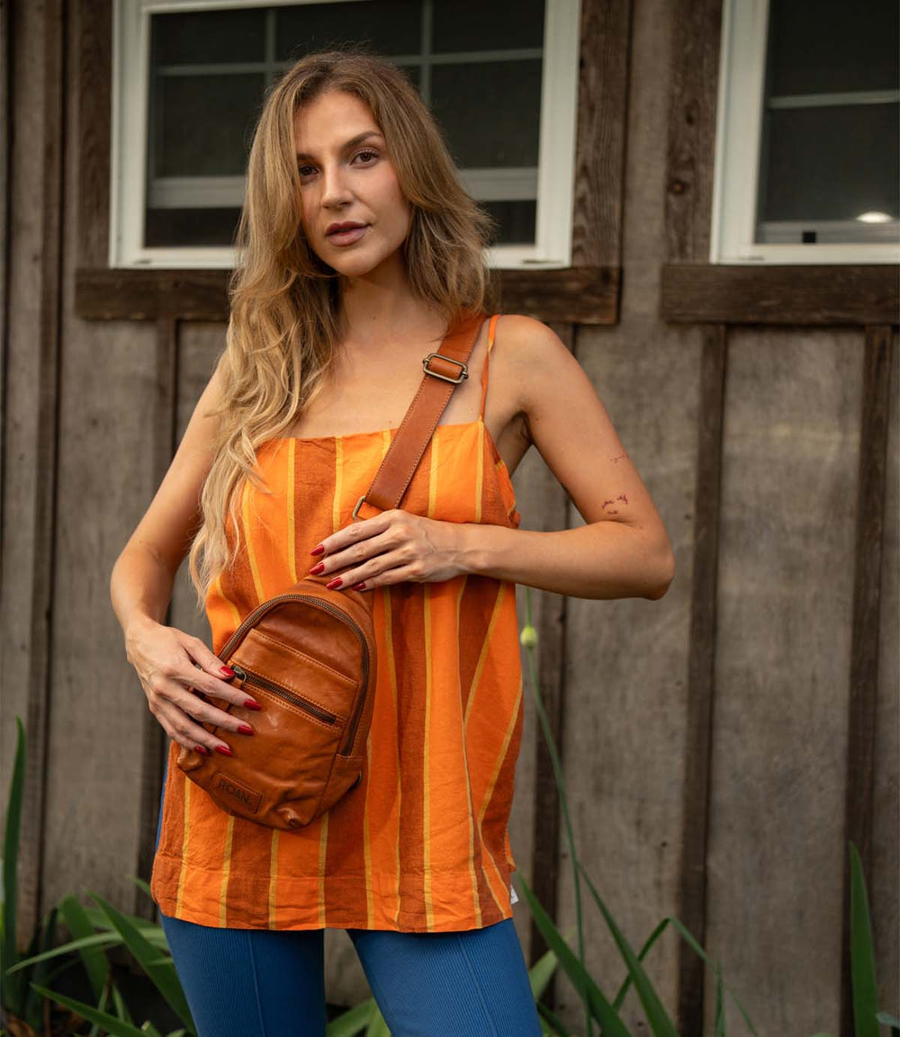 A woman with long blonde hair stands before a wooden building, wearing Roan's Trickle orange striped top, blue pants, and carrying a compact leather bag.